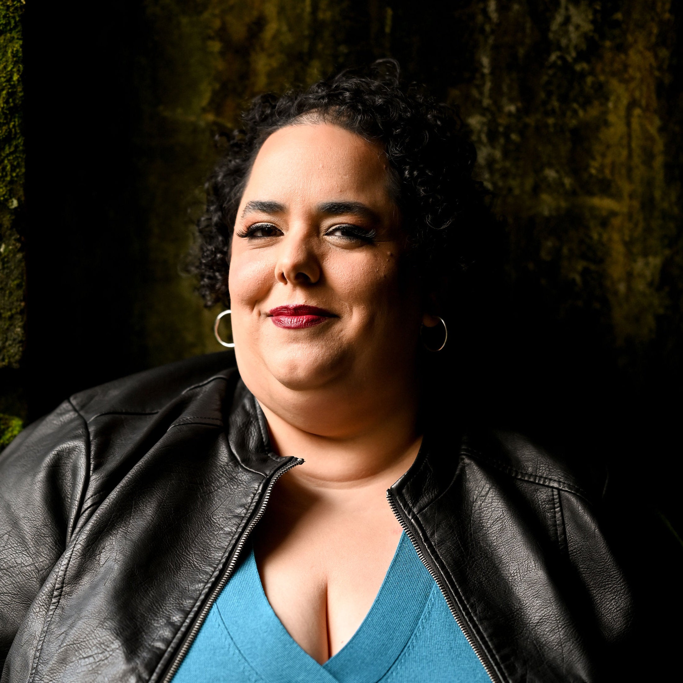 Photo of Coral Alejandra Moore, a woman with brown skin, curly black hair, and red lipstick wearing a turquoise shirt and a black leather jacket.